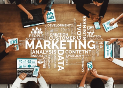 Top-down view of a diverse marketing team collaborating around a wooden table with laptops and tablets, overlaid with strategic keywords like 'Marketing', 'Data', 'Content', and 'Customer'.
