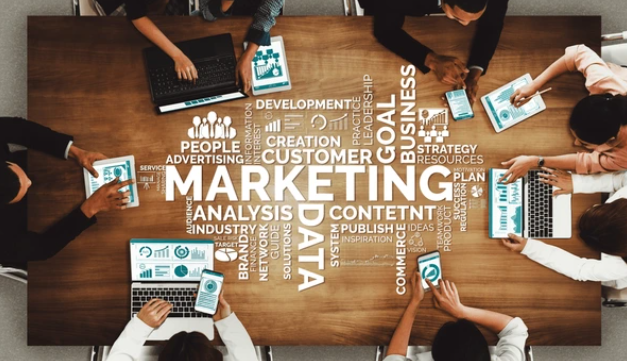 Top-down view of a diverse marketing team collaborating around a wooden table with laptops and tablets, overlaid with strategic keywords like 'Marketing', 'Data', 'Content', and 'Customer'.